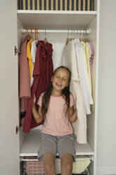 Glückliches Mädchen mit geschlossenen Augen sitzt bei Kleidung im Schrank - LESF00033