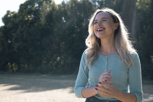 Fröhliche blonde Frau steht im Park an einem sonnigen Tag - AMWF00618