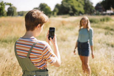 Nicht-binäre Person, die ihren Freund an einem sonnigen Tag mit dem Smartphone fotografiert - AMWF00628