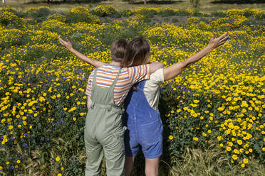 Non-binary person hugging friend standing with arms outstretched in front of flowering plants - AMWF00638