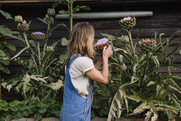 Mature woman holding flower standing in front of plant - AMWF00650