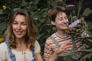 Lächelnde reife Frau mit Freund, der eine Blume betrachtet, die inmitten von Pflanzen steht - AMWF00651
