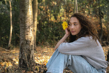 Beautiful young woman with yellow flower sitting at park - AMWF00662