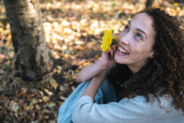 Glückliche junge Frau mit gelber Blume im Park sitzend - AMWF00664