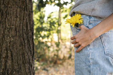 Woman with yellow flower in jeans pocket standing by tree - AMWF00669