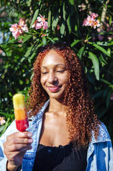 Lächelnde junge Frau mit Eiscreme vor einer Pflanze stehend - ASGF02797