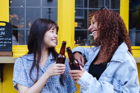 Glückliche Freunde stoßen mit Bierflaschen vor einem Fenster an - ASGF02803