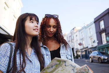 Smiling young woman with friend holding map standing on street - ASGF02815