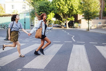 Cheerful friends holding hands and running on road in city - ASGF02826