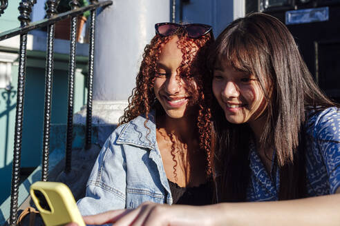Lächelnde junge Frau mit Freund bei der Nutzung eines Smartphones an einem sonnigen Tag - ASGF02829