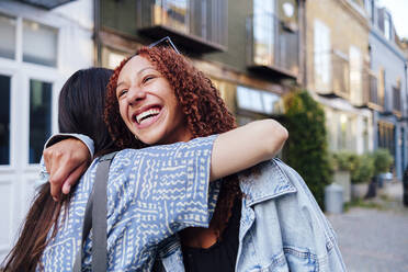 Happy woman with curly hair hugging friend in front of building - ASGF02836