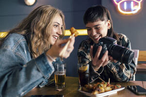 Smiling young lesbian couple with camera sitting at table in restaurant - JSRF02211