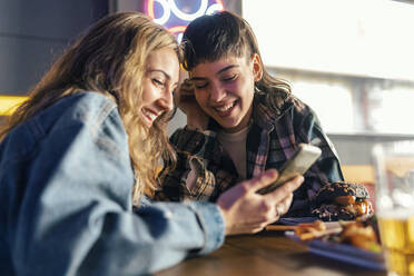 Fröhliches lesbisches Paar, das am Tisch im Restaurant ein Smartphone benutzt - JSRF02215