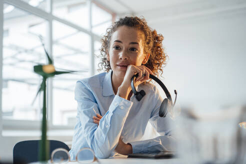 Young businesswoman holding headset looking at wind turbine in office - JOSEF12866