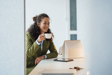 Happy businesswoman holding coffee cup sitting with model house on desk in office - JOSEF12871