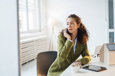 Young businesswoman talking on smart phone sitting with model home at desk in office - JOSEF12874