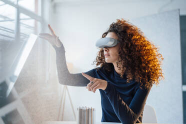 Young engineer with curly hair wearing virtual reality simulator sitting at office - JOSEF12889