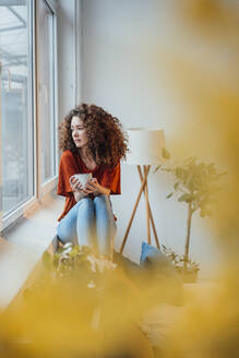 Junge Frau mit Kaffeetasse, die zu Hause am Fenster sitzt - JOSEF12913