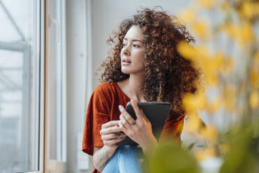 Thoughtful woman with tablet PC sitting by window at home - JOSEF12916