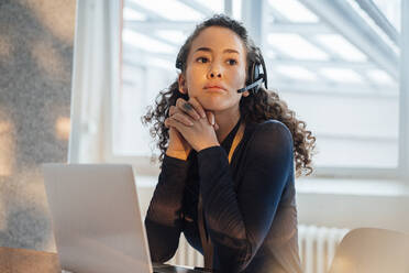 Young customer service representative sitting with hands clasped at desk in office - JOSEF12935