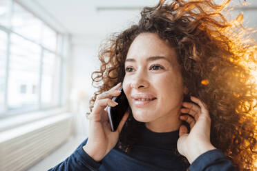 Lächelnde Frau mit lockigem Haar, die mit einem Smartphone telefoniert - JOSEF12975