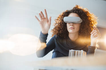 Geschäftsfrau mit lockigem Haar, die einen Virtual-Reality-Simulator trägt und im Büro gestikuliert - JOSEF12983