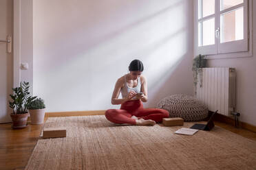 Junge Frau, die ihr Smartphone am Laptop benutzt und zu Hause auf dem Teppichboden sitzt - MMPF00241
