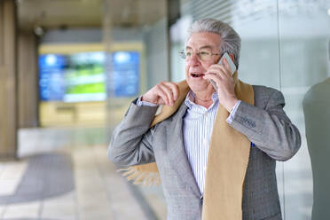 Senior businessman with scarf talking on smart phone standing by glass wall - GGGF01030
