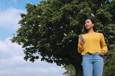 Nachdenkliche junge Frau mit Smartphone vor einem Baum stehend - AMWF00688