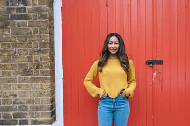 Smiling young woman with hands in pockets standing in front of red door - AMWF00705