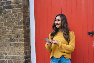 Glückliche junge Frau mit Handy vor einer roten Tür - AMWF00706