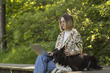 Junger Freiberufler arbeitet am Laptop mit süßem Hund im Park - SSGF00998