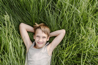 Smiling boy lying down with hands behind head on grass - EYAF02133
