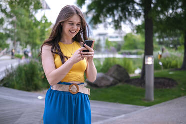 Happy young woman text messaging through mobile phone in park - AMWF00734