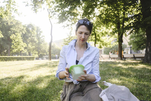 Frau mit Lunchpaket sitzt im Park an einem sonnigen Tag - EYAF02152