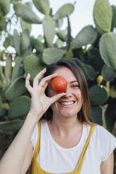 Smiling young farm worker covering eye with tomato - EGHF00540