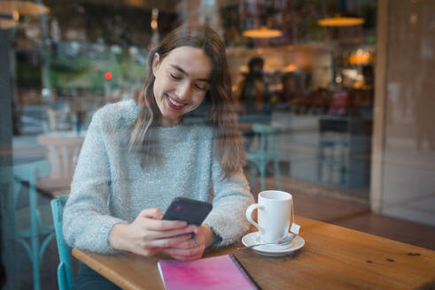 Inhalt Charmante Frau sitzt am Tisch im Café und surft lächelnd auf ihrem Smartphone - ADSF36300