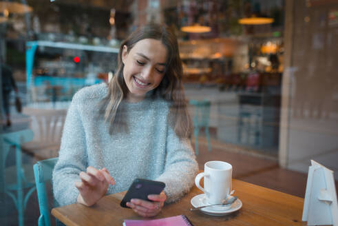 Inhalt Charmante Frau sitzt am Tisch im Café und surft lächelnd auf ihrem Smartphone - ADSF36301