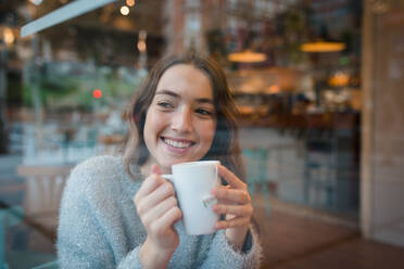 Glückliche Frau sitzt am Tisch im Café und trinkt ein leckeres Getränk, während sie aus dem Fenster schaut und das Wochenende genießt - ADSF36303