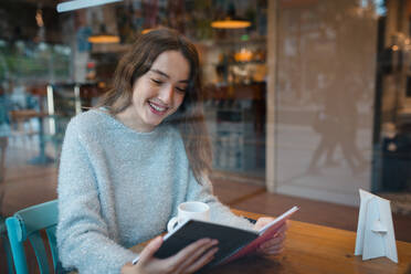 Positive junge Frau sitzt am Tisch in einem Café und genießt ein interessantes Buch, während sie lächelt und einen schönen Tag genießt - ADSF36306