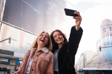 Low angle of cheerful female friends standing on street in Madrid and taking self shot on smartphone during city stroll - ADSF36307