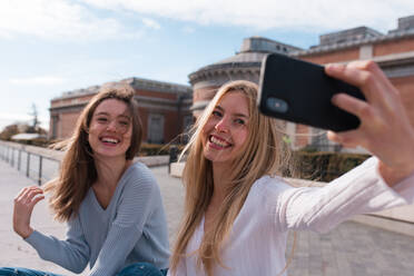 Cheerful female friends standing on street in Madrid and taking self shot on smartphone during city stroll - ADSF36309