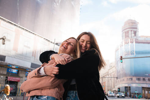 Low angle of smiling female best friends cuddling on street in Madrid and enjoying weekend together - ADSF36310