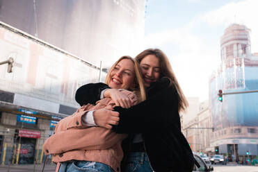 Low angle of smiling female best friends cuddling on street in Madrid and enjoying weekend together - ADSF36311