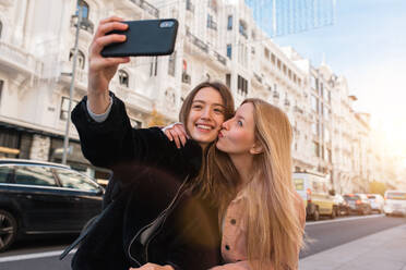 Content best female friends standing on street and taking selfie on smartphone while enjoying weekend together in Madrid - ADSF36314