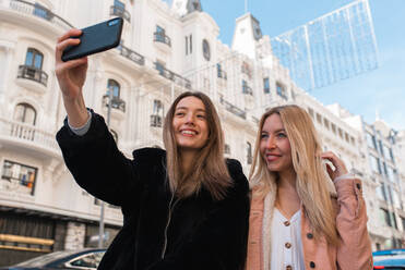 Content best female friends standing on street and taking selfie on smartphone while enjoying weekend together in Madrid - ADSF36315
