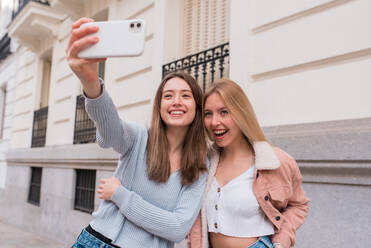 Content best female friends standing on street and taking selfie on smartphone while enjoying weekend together in Madrid - ADSF36316