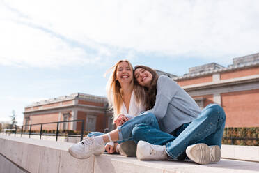 Content female friends in stylish clothes sitting on street in Madrid and hugging while looking at camera and spending time together at weekend - ADSF36320