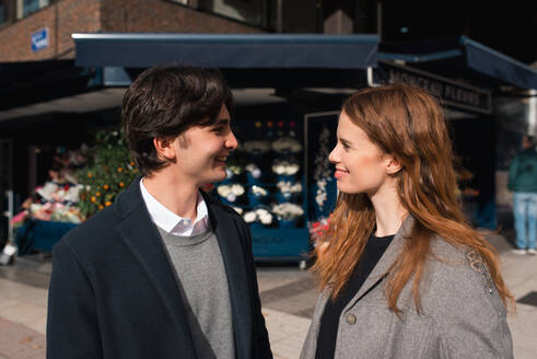 Seitenansicht eines lächelnden jungen Paares, das während eines romantischen Rendezvous in der Stadt auf einer Straße zusammensteht - ADSF36322