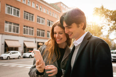 Happy young man standing with girlfriend while browsing on smartphone on the city street - ADSF36327